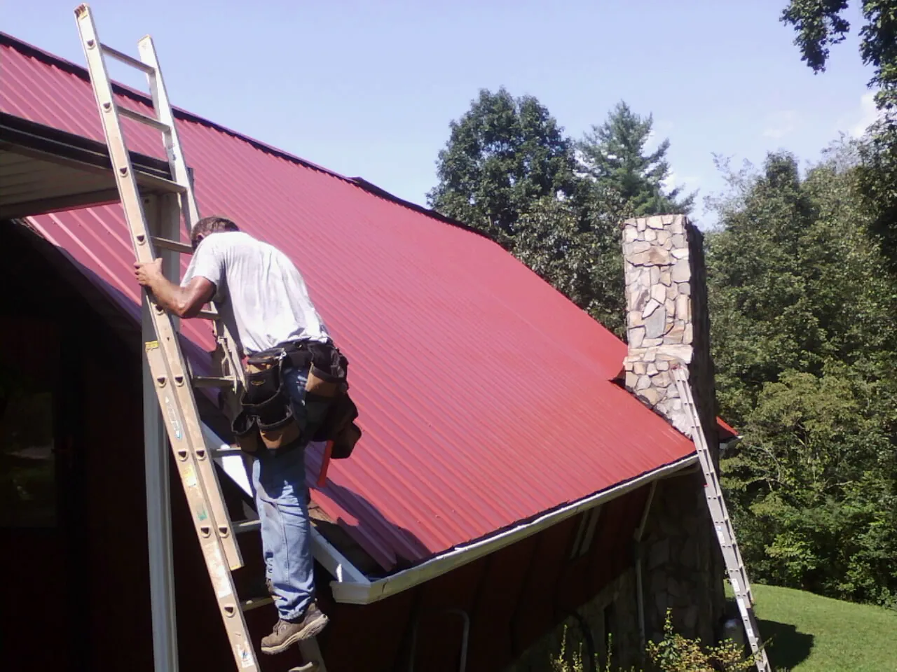 Roofer on a ladder beside a red metal roof and stone chimney — Soffit & Fascia Repair by Roofing Brothers in Crandall